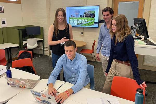 Four students collaborating around a laptop in a university classroom, with a presentation on 'Forced Labor in Supply Chains' displayed on a screen in the background.