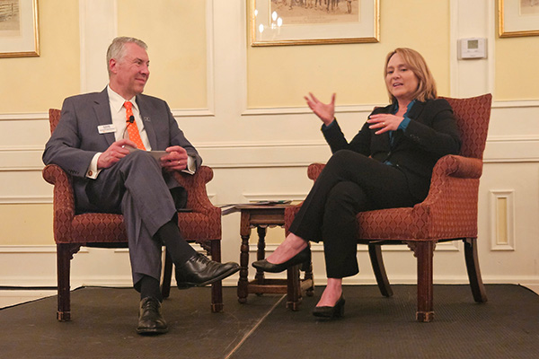 Two people seated in armchairs are engaged in conversation on a stage. The person on the left is holding papers, while the person on the right is gesturing with hands. The setting appears to be a formal event. The room has framed pictures on the walls.