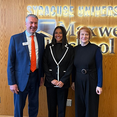 Three individuals are standing in front of a wooden wall with illuminated signage that reads "Syracuse University Maxwell School." They are dressed in formal attire.