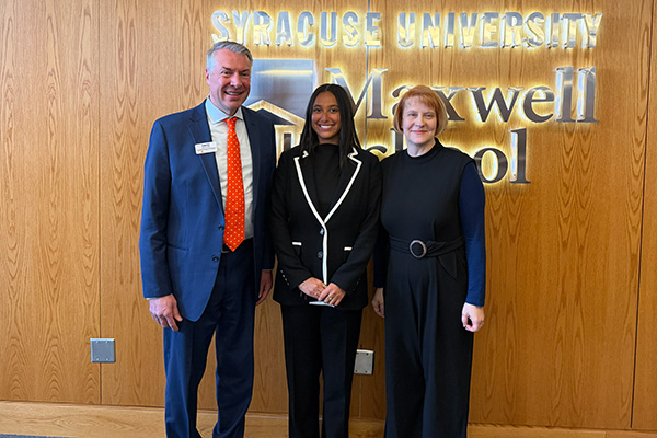 Three individuals are standing in front of a wooden wall with illuminated signage that reads