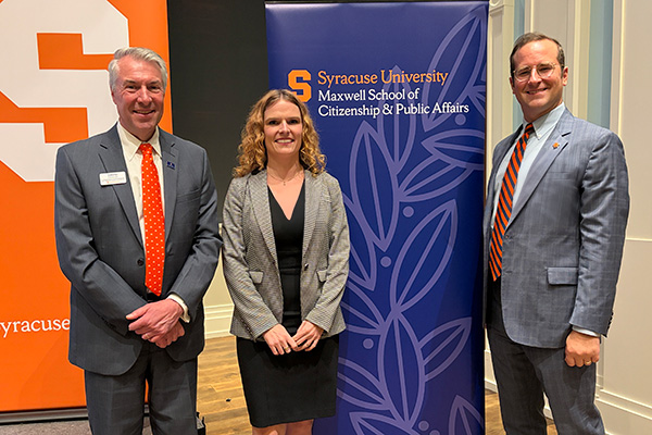Three people posing together near Maxwell School banner at an event.