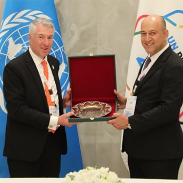 Two individuals are standing behind a table at a United Nations event, holding a decorative plate between them. There's a UN logo to the left and a Red Crescent logo to the right in the background.
