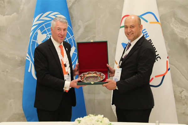 Two individuals are standing behind a table at a United Nations event, holding a decorative plate between them. There's a UN logo to the left and a Red Crescent logo to the right in the background.