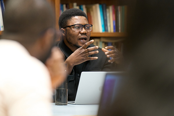 A person wearing glasses gestures while speaking during a seminar discussion, seated at a table with an open laptop, with bookshelves visible in the background.