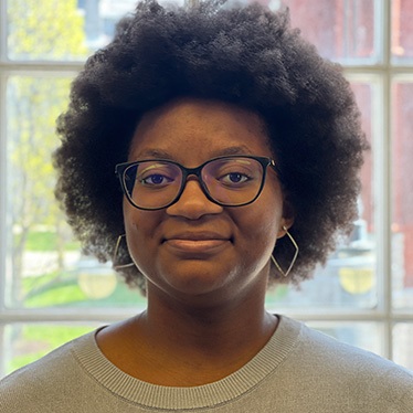 Portrait of an individual wearing glasses and a grey sweater, smiling in front of a window with a view of trees.