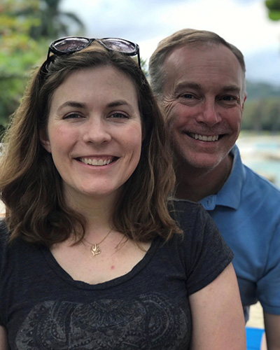 A person with long brown hair and sunglasses resting on their head smiles in the foreground, with another person in a light blue polo shirt smiling behind them, with a beach and trees visible in the background.