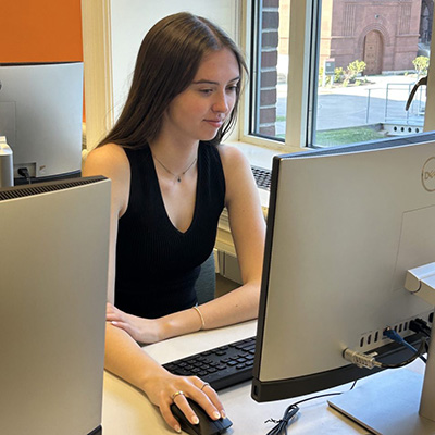 A person works at a desktop computer in an office, with a window overlooking a brick building in the background.