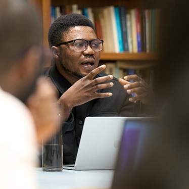 A person wearing glasses gestures while speaking during a seminar discussion, seated at a table with an open laptop, with bookshelves visible in the background.
