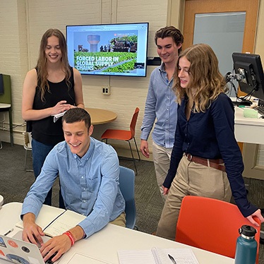 Four students collaborating around a laptop in a university classroom, with a presentation on 'Forced Labor in Supply Chains' displayed on a screen in the background.