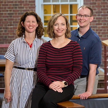 Three individuals, smiling, in front of a brick building with windows. The person in the center is seated, while the other two stand beside them.