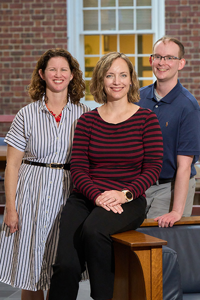 Three individuals, smiling, in front of a brick building with windows. The person in the center is seated, while the other two stand beside them.