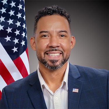A person in a suit smiles in front of a U.S. flag.