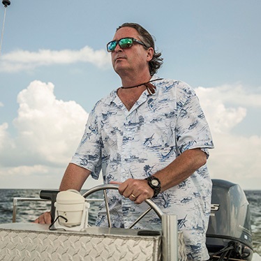 A person is steering a boat on open water, wearing a patterned shirt and sunglasses, with a cloud-filled sky in the background.