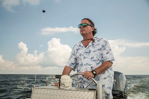 A person is steering a boat on open water, wearing a patterned shirt and sunglasses, with a cloud-filled sky in the background.