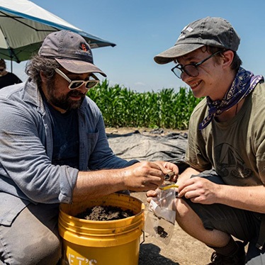 Two people crouch over a bucket, examining soil samples at an outdoor excavation site.