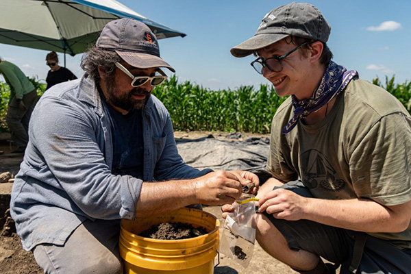 Two people crouch over a bucket, examining soil samples at an outdoor excavation site.