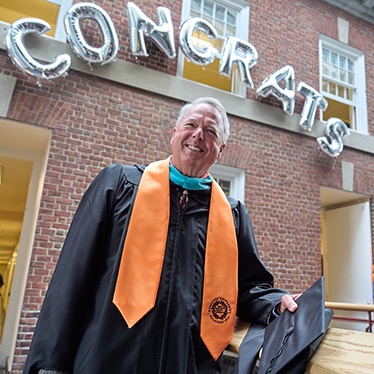 An individual in graduation attire stands smiling in front of a brick building. Above them, silver balloon letters spell "CONGRATS." Balloon decorations in orange, blue, and white are visible in the background.