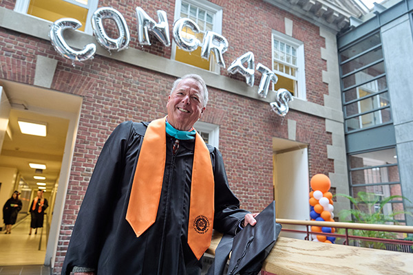 An individual in graduation attire stands smiling in front of a brick building. Above them, silver balloon letters spell