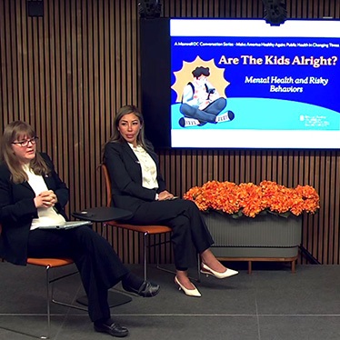 A panel discussion setup with two individuals seated on chairs in front of an audience. Behind them is a screen displaying a slide titled "Are The Kids Alright? Mental Health and Risky Behaviors."