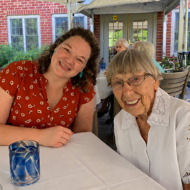 Two people smiling at an outdoor table, one younger with curly hair, one older with glasses.