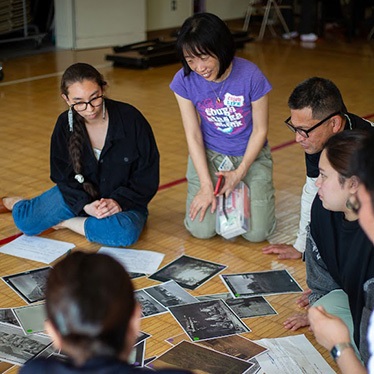 A group of individuals is seated on the floor, gathered around a collection of photographs. They are engaged in discussion, with one person holding a notebook and another wearing glasses. The setting appears informal and collaborative.