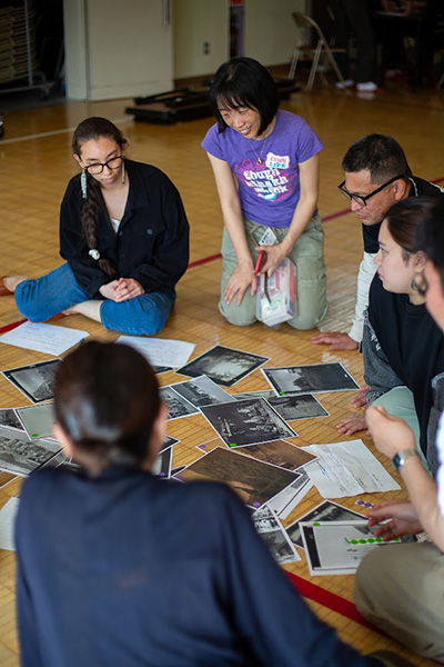 A group of individuals is seated on the floor, gathered around a collection of photographs. They are engaged in discussion, with one person holding a notebook and another wearing glasses. The setting appears informal and collaborative.
