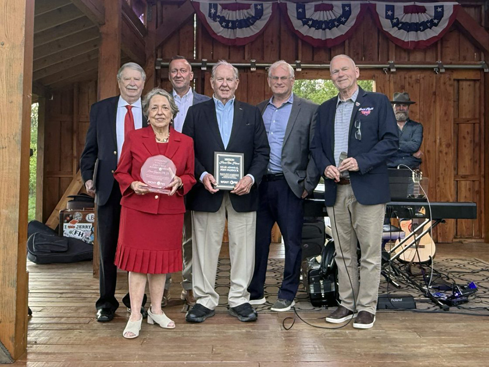 A group of six people stands on a wooden stage. Some are holding plaques and an award. Musical instruments are visible in the background. The stage is decorated with patriotic bunting.