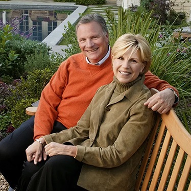 Two smiling people, one in an orange sweater and one in a tan jacket, sit together on a garden bench, with a pool and landscaped yard behind them.