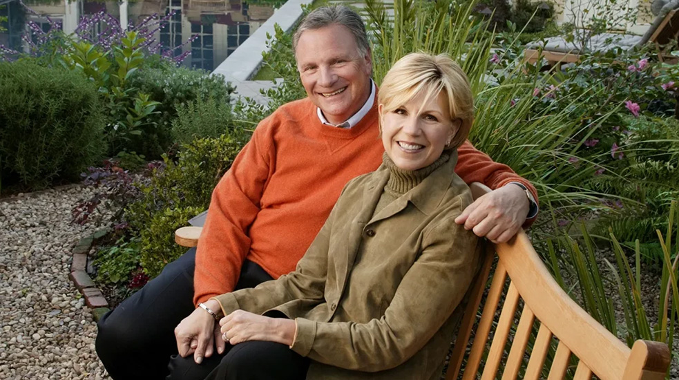 Two smiling people, one in an orange sweater and one in a tan jacket, sit together on a garden bench, with a pool and landscaped yard behind them.
