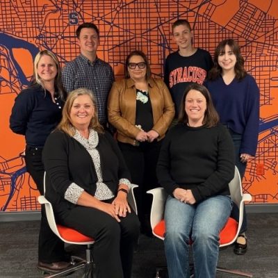 Group of seven people posing for a photo in a room with a bright orange map of Syracuse in the background.