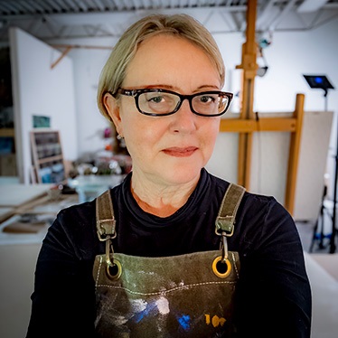 A person wearing black-rimmed glasses and a paint-splattered apron stands in an art studio, with an easel visible in the background.