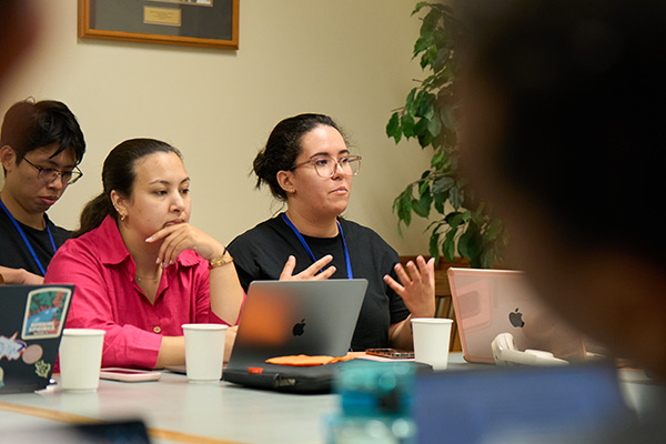 Three seminar participants seated at a table with laptops and coffee cups, one gesturing with both hands while speaking, others listening attentively.