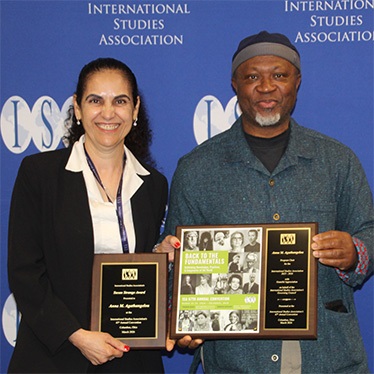 Two individuals stand in front of an "International Studies Association" backdrop. One is holding two plaques, and the other is holding a large framed recognition item with "Back to the Fundamentals" prominently displayed.