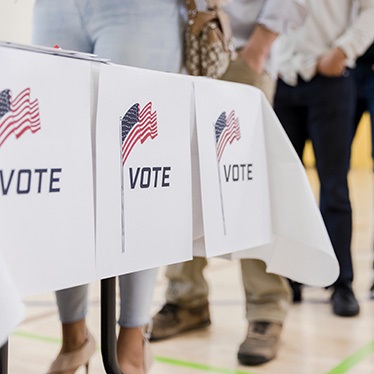 Voters stand in line at a table with “Vote” signs featuring American flags at a polling site.
