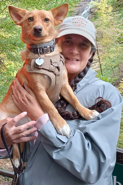 A person in a rain jacket holds a small tan dog in front of a waterfall.