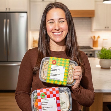 A person smiles and holds two packaged prepared meals in a kitchen setting. The meal containers are labeled "Spaghetti and Meatballs" and "Mac and Cheese," with bilingual packaging in English and Spanish.