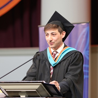 Male student in graduation regalia speaking at a podium