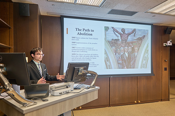 A person is giving a presentation in a lecture room. The slide on the screen is titled