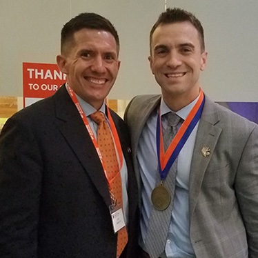 Two individuals smiling at the camera, one wearing a medal, at an indoor event with a "THANK YOU TO OUR SPONSORS" sign in the background.