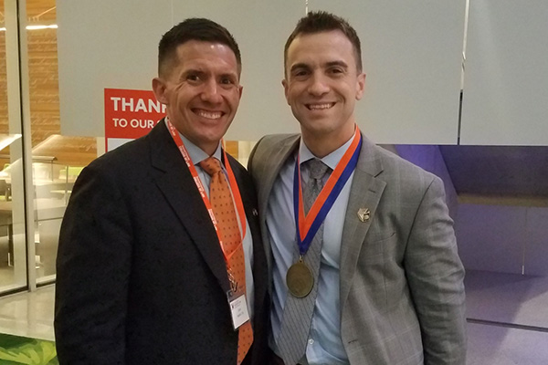 Two individuals smiling at the camera, one wearing a medal, at an indoor event with a