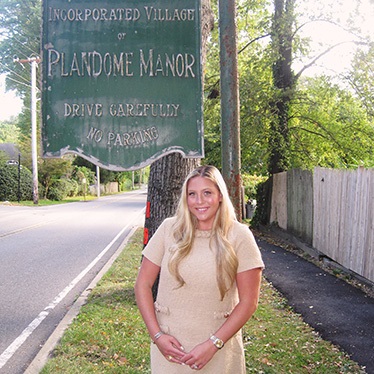 A person smiling, standing in front of the "Incorporated Village of Plandome Manor" sign on a street.