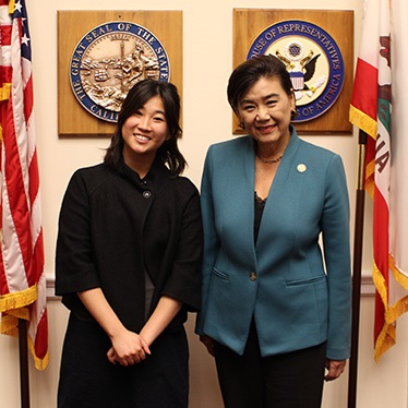 wo individuals stand smiling in an office setting between the flags of the United States and California. Behind them are plaques with official seals.