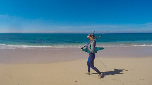 Person walking on a sandy beach carrying a spear gun. The ocean is calm under a clear blue sky.