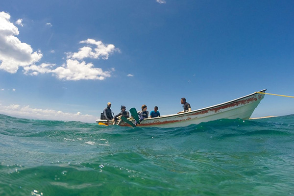 A group of people is seated in a small boat on the ocean under a clear blue sky. The water is calm, with gentle waves. There are a few clouds in the sky.