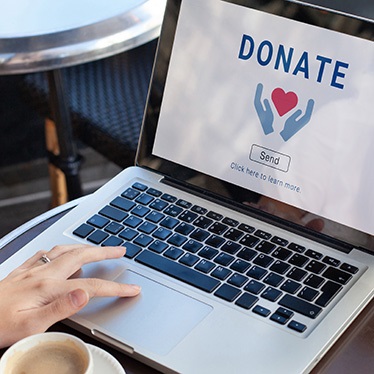 A person seated at a round table is using a laptop. The screen displays the word "DONATE" above an illustration of hands holding a heart, with a "Send" button below.