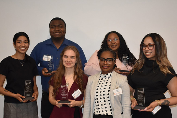 A group of six individuals smiles while holding awards in front of a plain background.