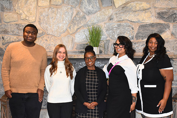 Group of five individuals smiling and standing in front of a stone wall. Two are wearing sweaters, one in professional attire, one in a white shirt, and one in a chef's uniform.