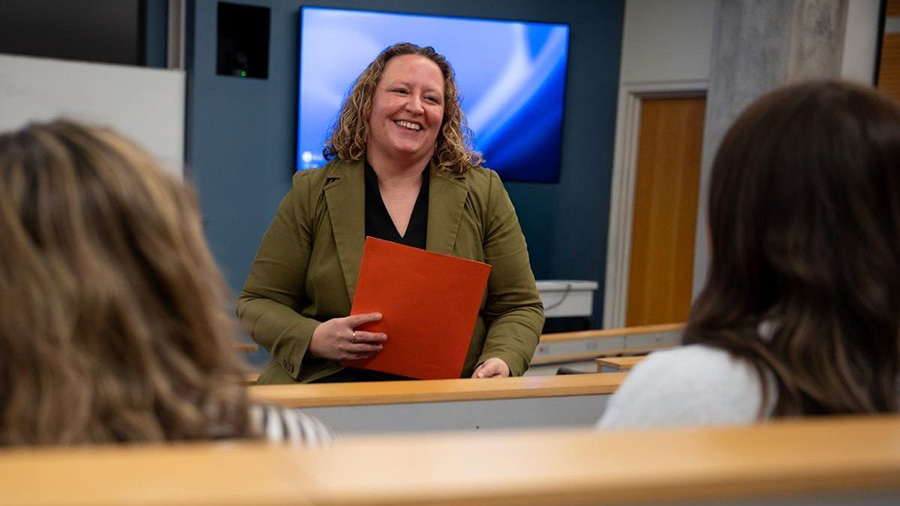 Instructor smiling while teaching, holding a red folder in a classroom with students.