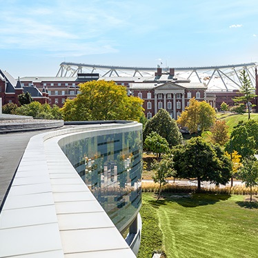 Exterior view of a modern curved building with reflective windows in the foreground, overlooking an area with lush greenery, with the traditional red brick architecture of the Maxwell School visible in the background under a clear blue sky.