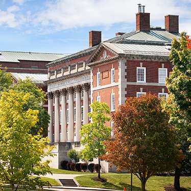View of Maxwell Hall on a sunny day, featuring its classic red brick facade and large white columns, surrounded by green trees and a manicured lawn.